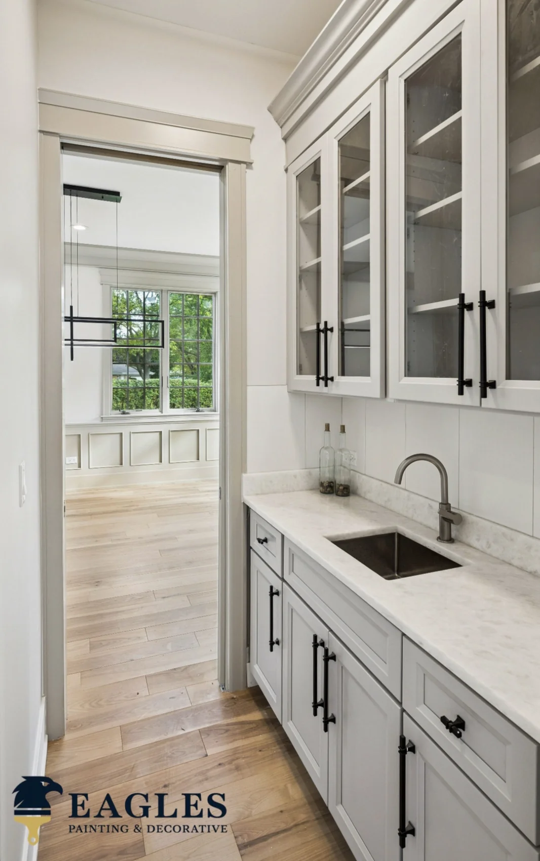 After cabinet refinishing showing a completed light gray wet bar with glass front uppers, black hardware, white counter, and stainless sink
