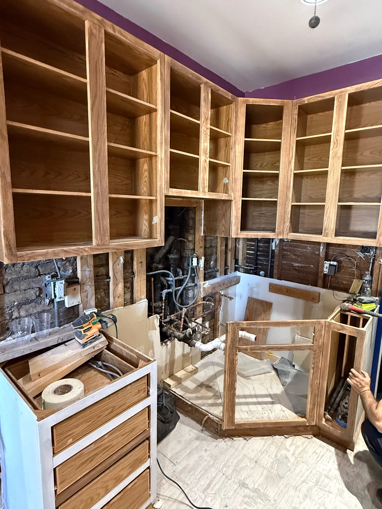 Before cabinet refinishing with doors removed, bare wood surfaces, exposed utilities, tools, and a protected work area