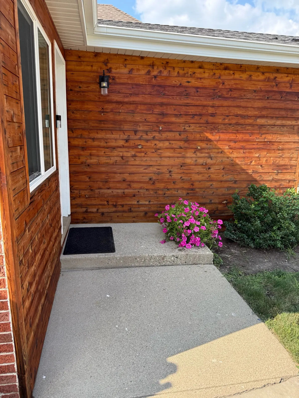 Residential entry with warm horizontal wood siding, white window trim, exterior light, and flowering landscape