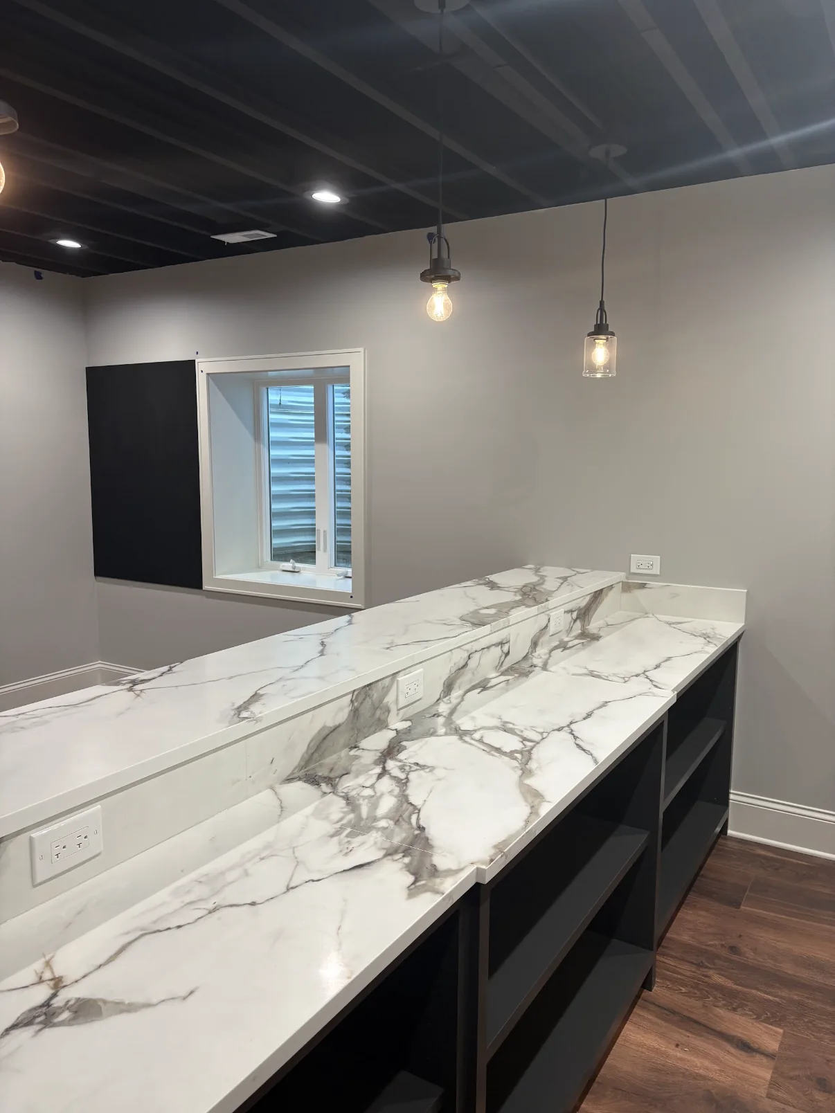 Finished interior bar room with light gray walls, dark open ceiling, marble look counters, and black built-in shelving