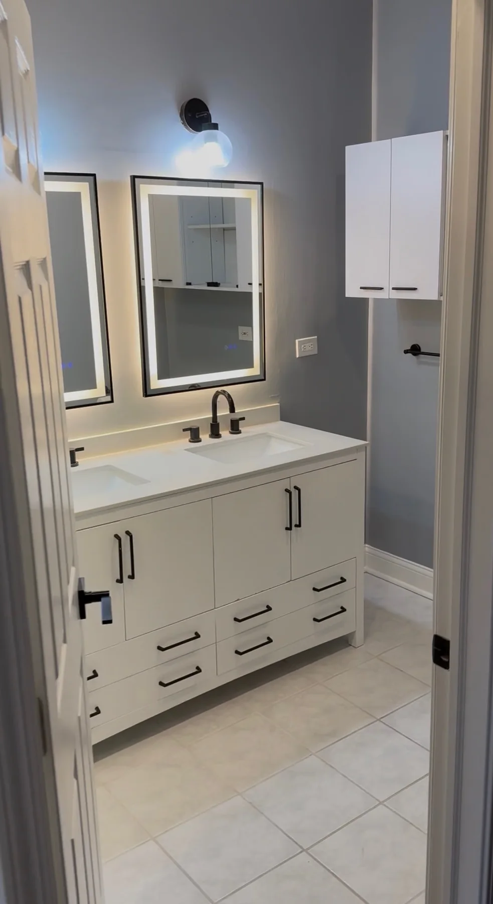 Finished bathroom with gray walls, white double vanity, framed mirrors, and upper cabinets
