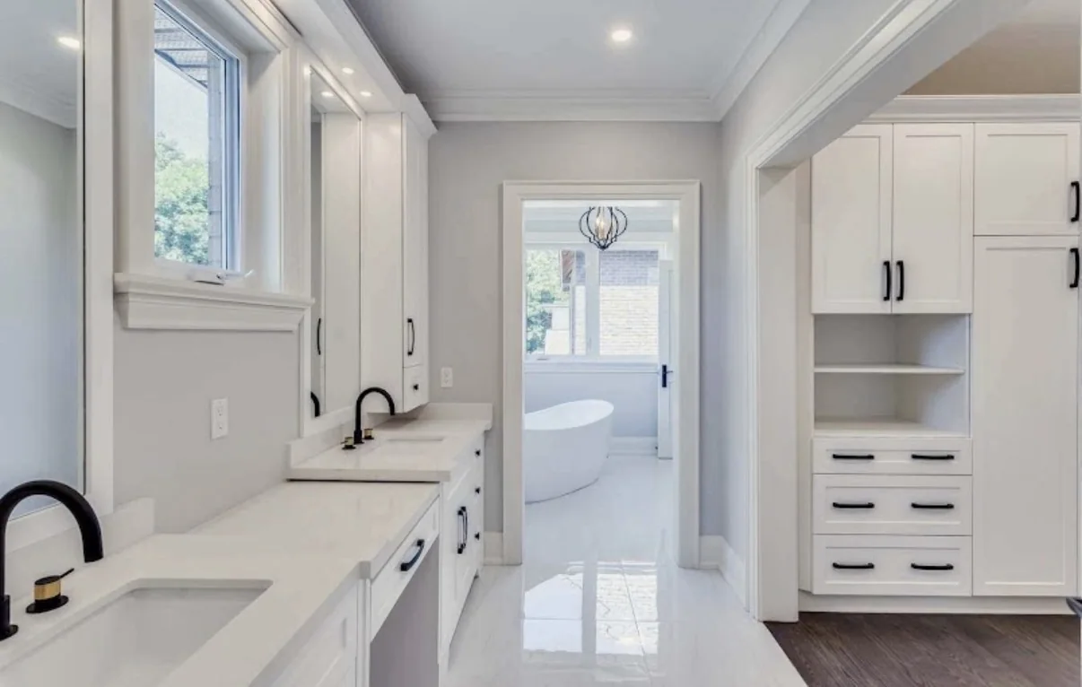 Bright interior with light gray walls, white cabinetry, double sinks, and a doorway to an adjoining bathroom