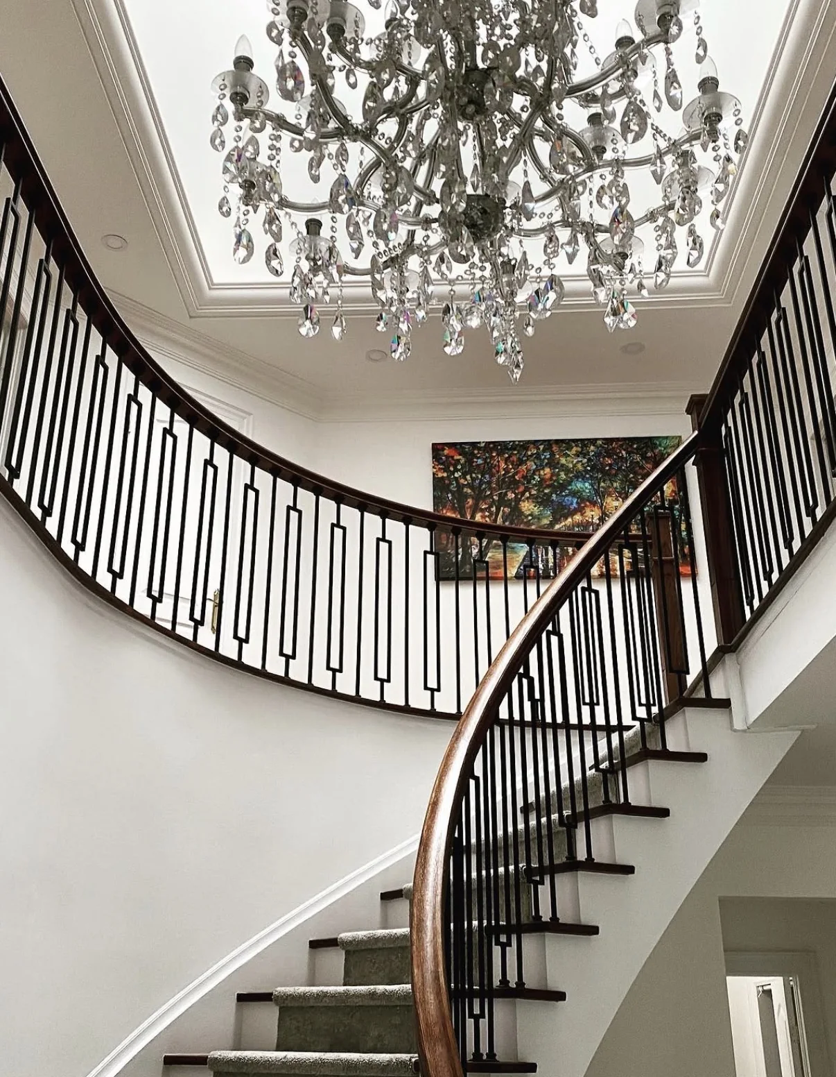 Curved residential staircase with white walls, dark wood handrail, geometric balusters, and a crystal chandelier