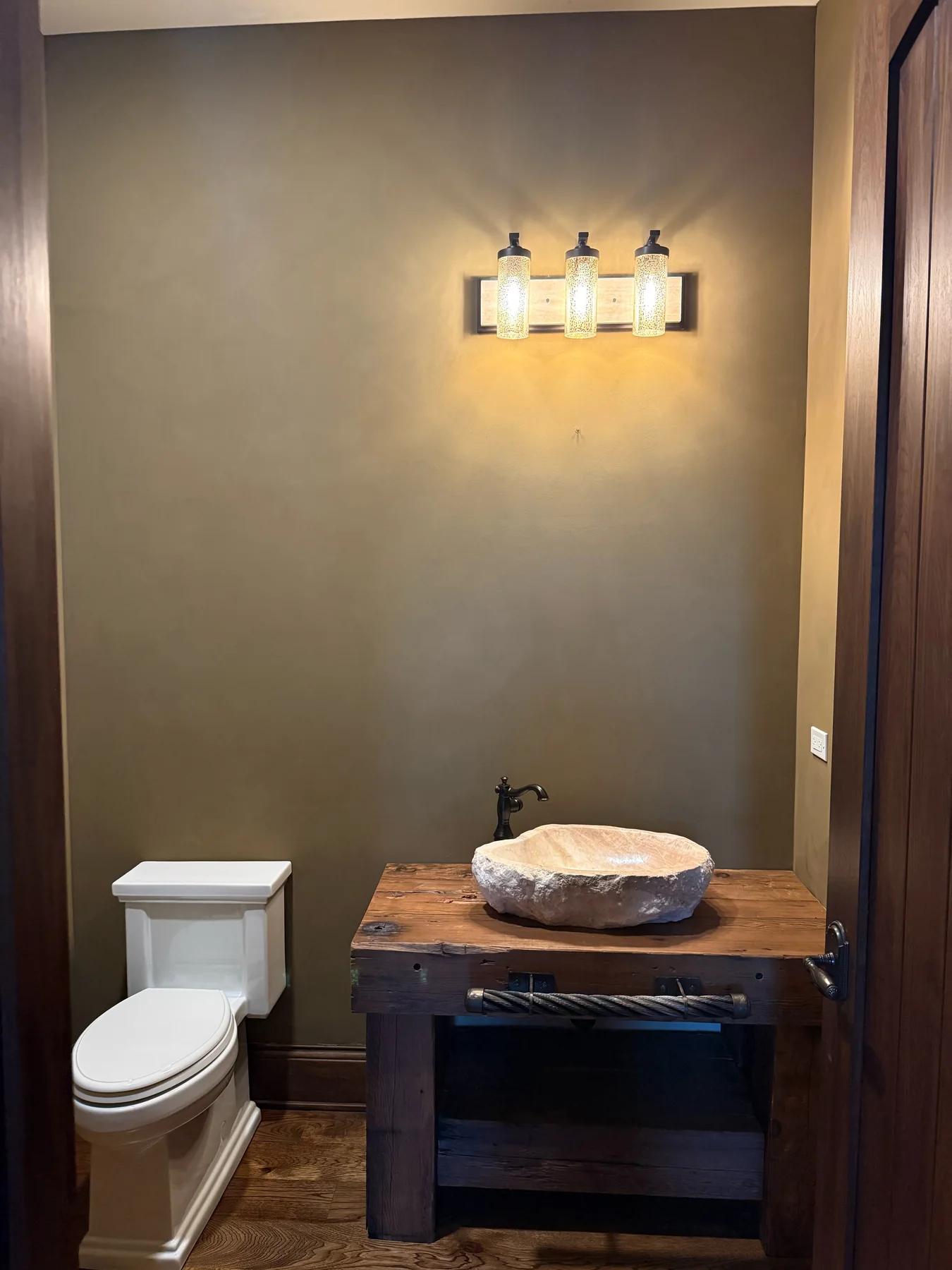 Completed powder room with warm limewash walls, wood vanity, stone vessel sink, wall lighting, and white toilet