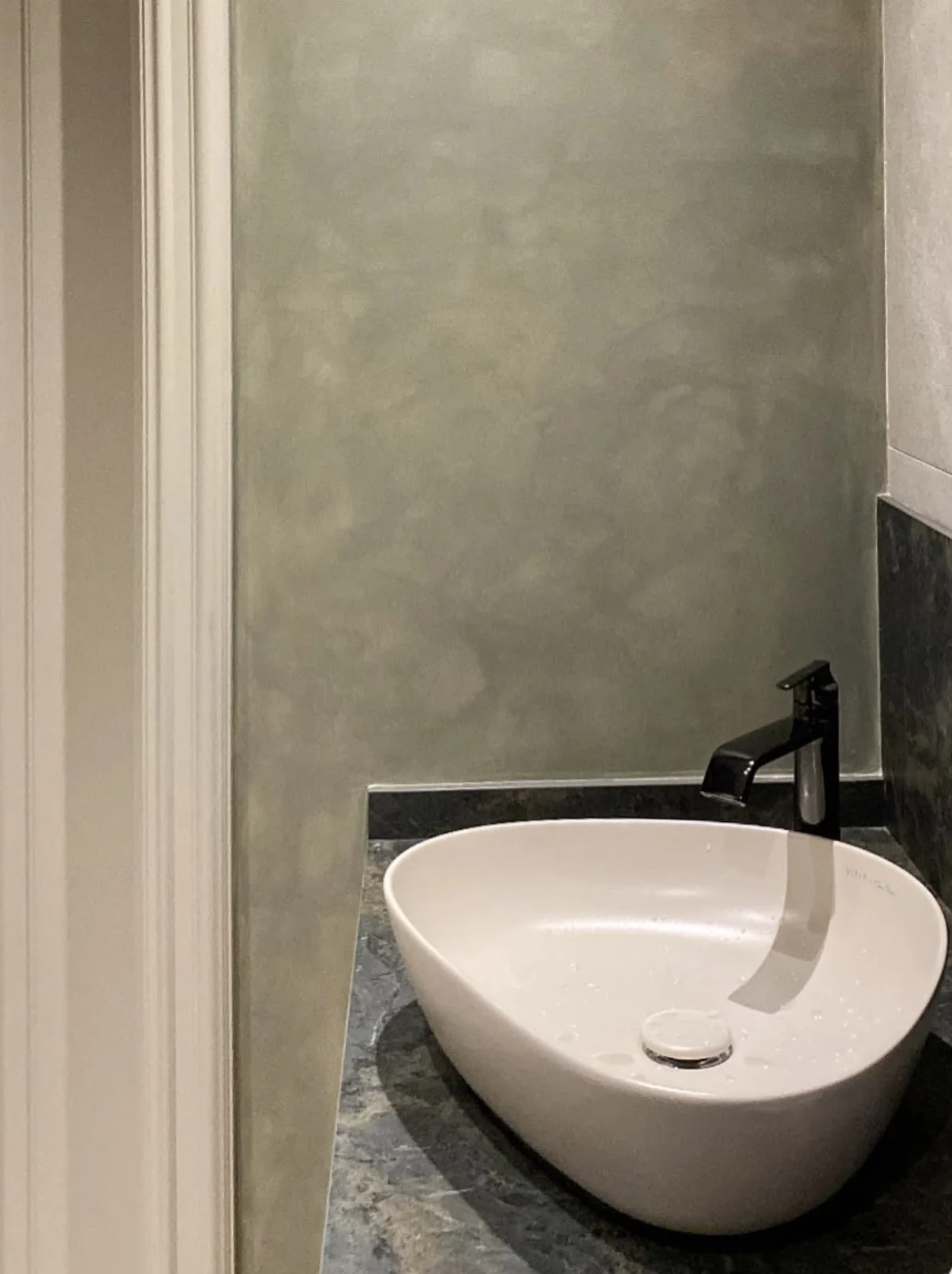 Detail of sage green limewash wall above a white vessel sink with a black faucet and dark stone vanity