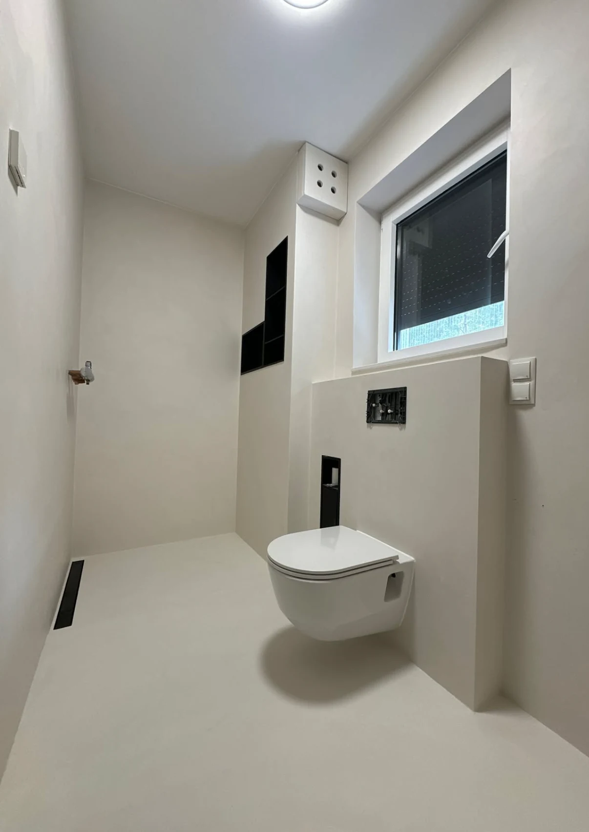 Pale microcement bathroom surfaces around a wall hung toilet, window and recessed storage