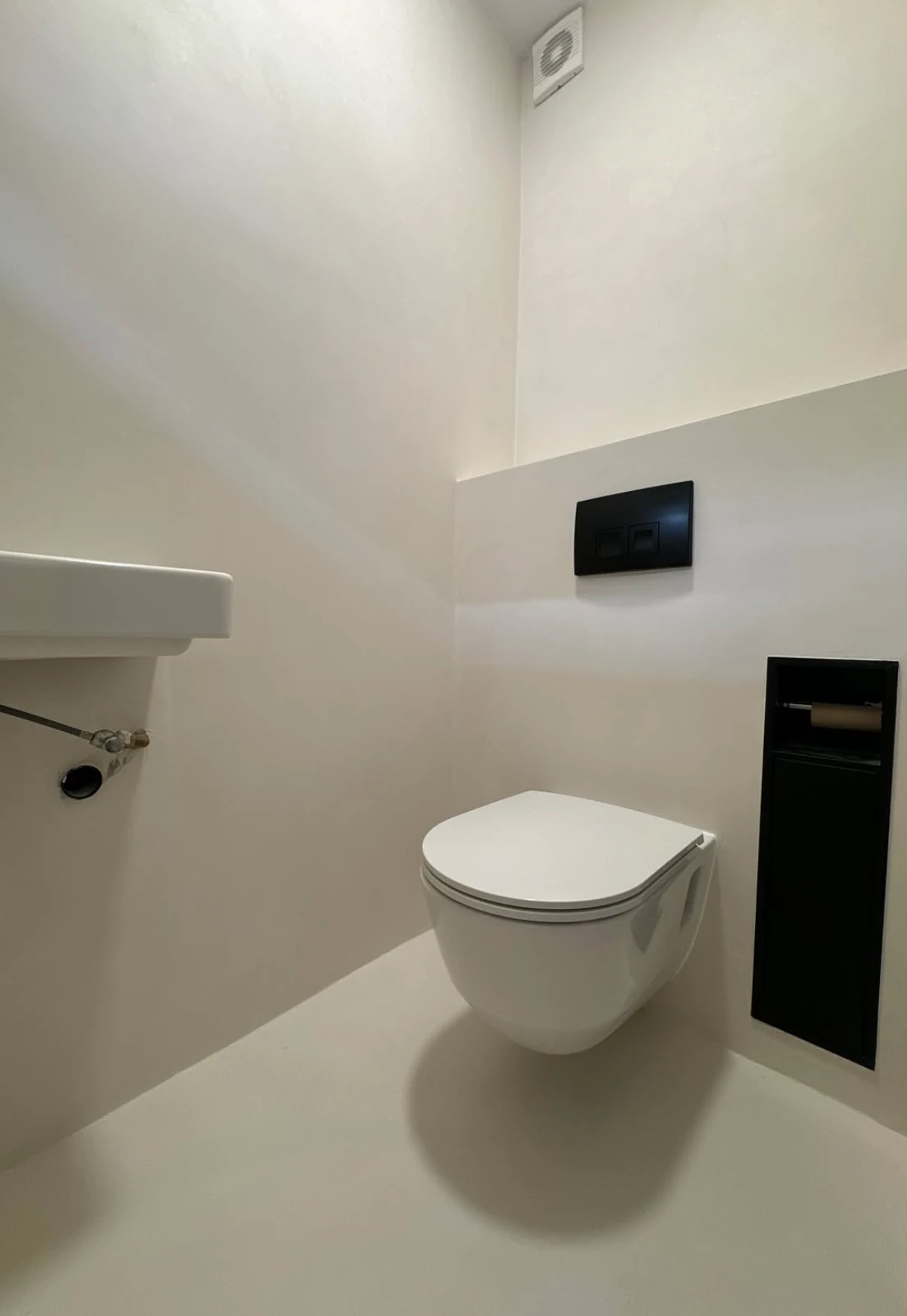 Light microcement wall and floor surfaces beside a wall hung toilet and black recessed fittings