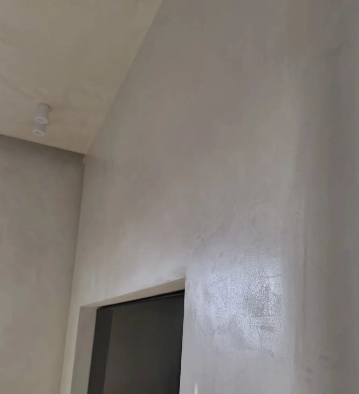 Warm gray microcement wall with mottled texture and soft sheen beside a dark interior doorway