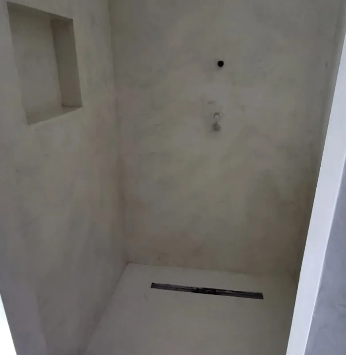 Walk-in shower installation in progress with pale continuous coated walls, a recessed niche, open fixture points, and a linear drain