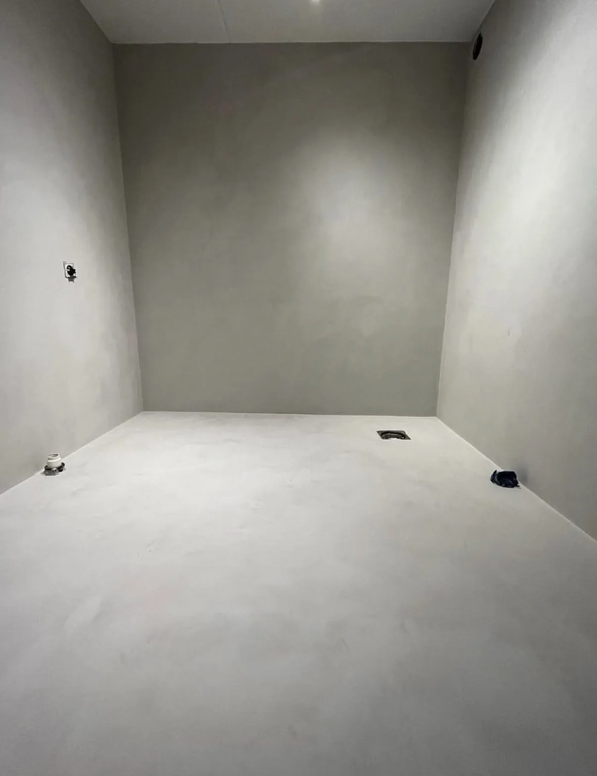 Wet-room installation in progress with seamless gray coated walls, pale floor, open drain, and exposed service points