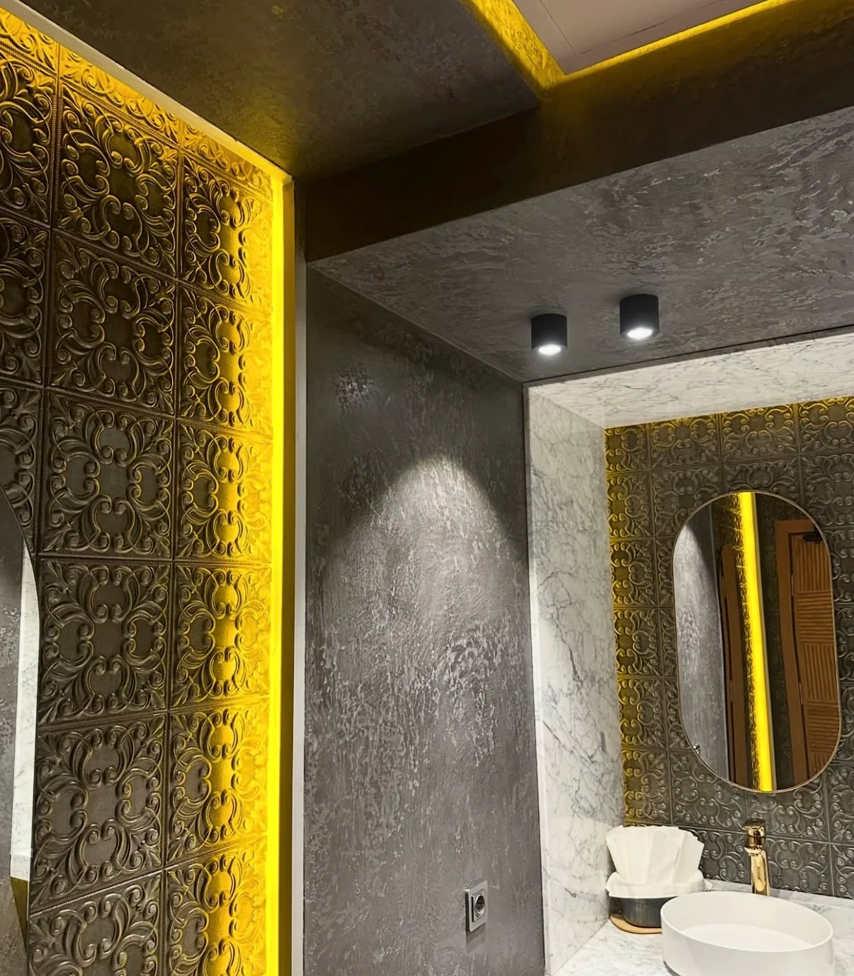 Textured gray Venetian plaster beside an ornate panel and oval mirror with warm accent lighting