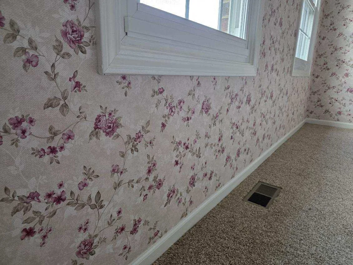 Finished floral wallpaper across a carpeted room, wrapping a corner and continuing around two white trimmed windows