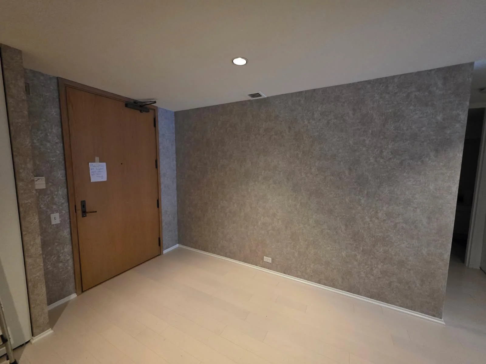 Finished warm gray taupe textured wallpaper across an empty room and narrow return beside a wood door
