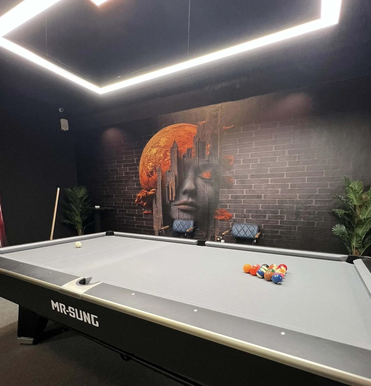 Dark mural of a stylized woman's face and orange moon installed behind a pool table in a billiards room
