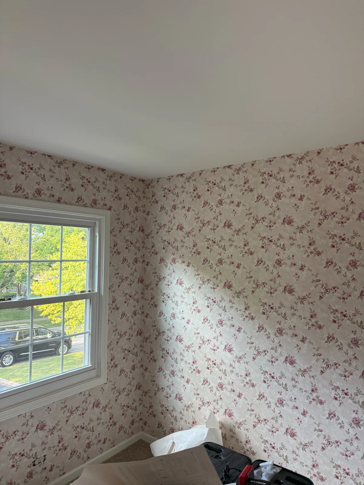 Floral wallpaper installed around a bright room corner and window while tools and packing materials remain on the floor