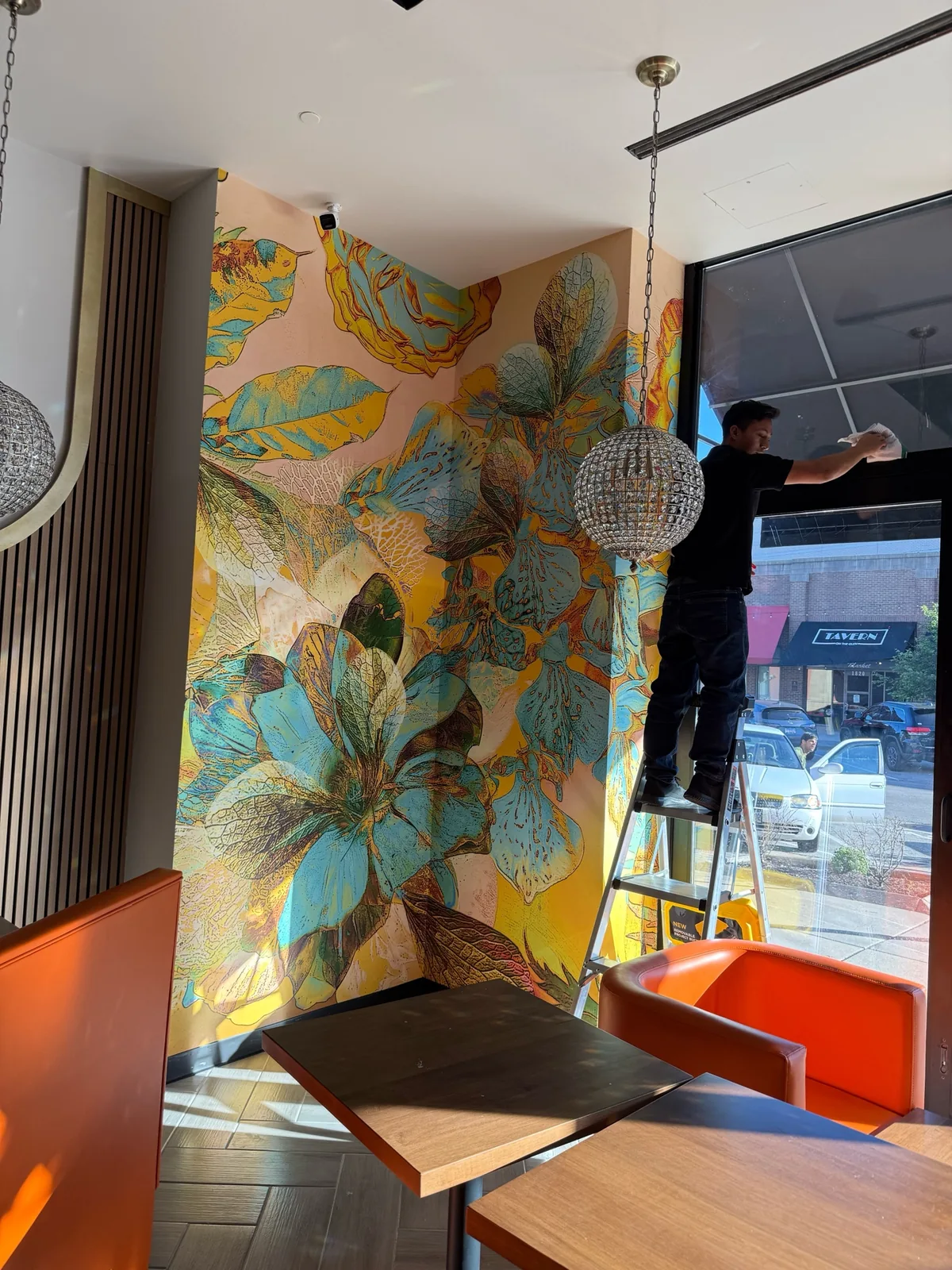Wallpaper installer on a step ladder smoothing an oversized floral wallcovering beside a restaurant window
