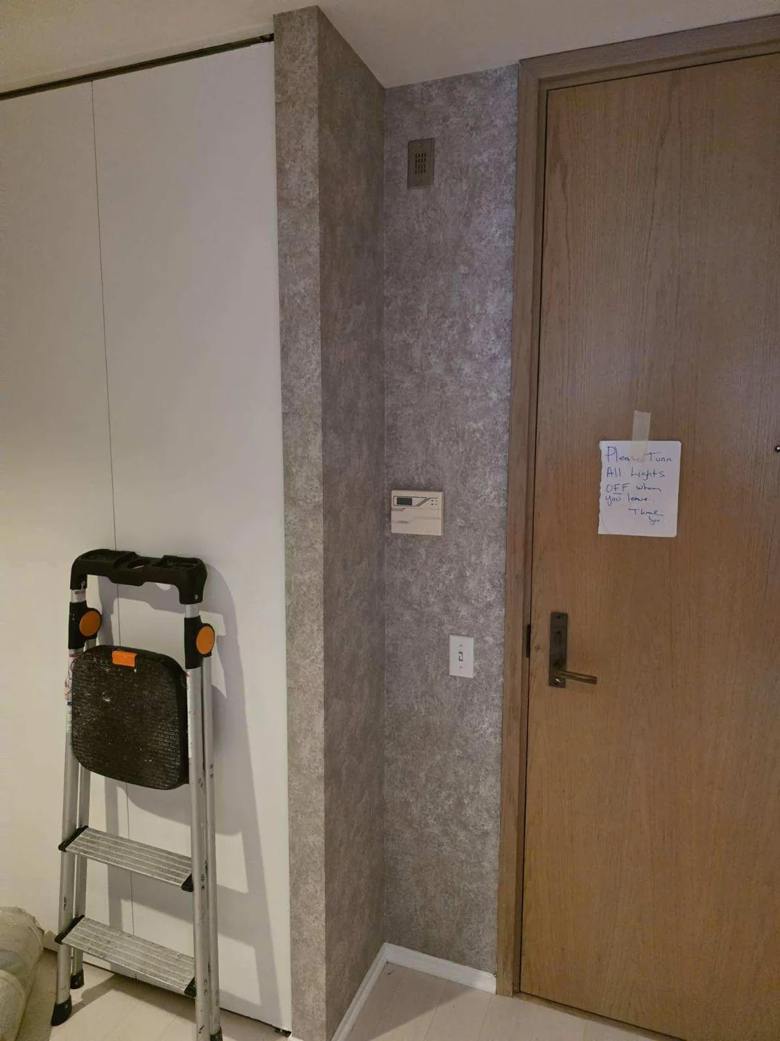 Gray textured wallpaper installed around a narrow entry corner, thermostat, outlet, and wood door beside a step ladder